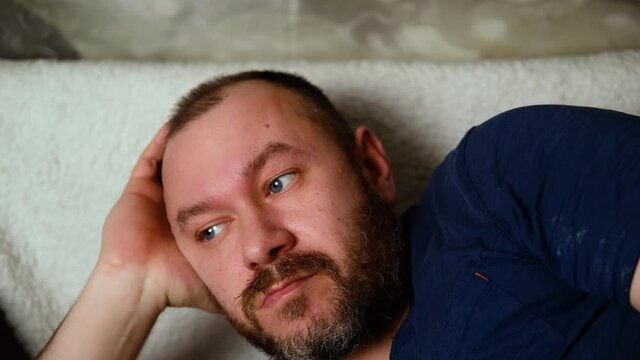 A tired, unshaven adult man lies on the sofa with a sleepy look.