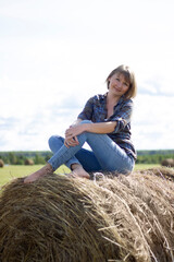 Naklejka na ściany i meble Beautiful modern young woman wearing in blue jeans and a plaid shirt posing against a haystack in field. 