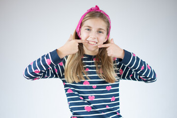Cute little girl smiles pointing fingers at mouth over white background.