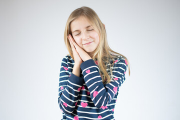 Portrait of cute little sleepy girl stands over white background with closed eyes and makes sleeping gesture.
