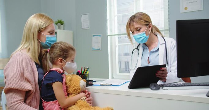 Medium Shot Of Female Pediatrician With Computer In Medical Mask Showing Tablet To Mother And Kid With Teddy Bear At Medical Visit In Hospital. Parent With Child At Doctor Office. Healthcare Concept