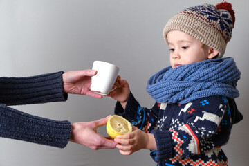 Sick child in clothes, toddler boy is standing on the grey background with a fever. llness child. Mother gives his son a lemon and a cup