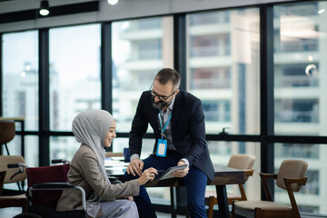 Friendly european executive officer is holding the tablet to show the information to muslim female officer. She is sitting in the wheelchair while she is discussing with her leader.