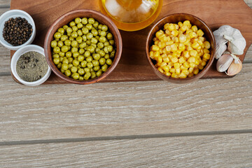 Bowls of boiled sweet corn and green peas, spices, oil, and vegetables on a wooden board