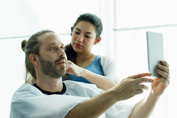 Fototapeta premium Family haircut at home, Beautiful wife is cutting her husband's hair during the quarantine lock down. He is holding a hand mirror to check his hairstyle.