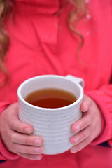 A white cup with tea in the hands of a girl in a pink jacket.