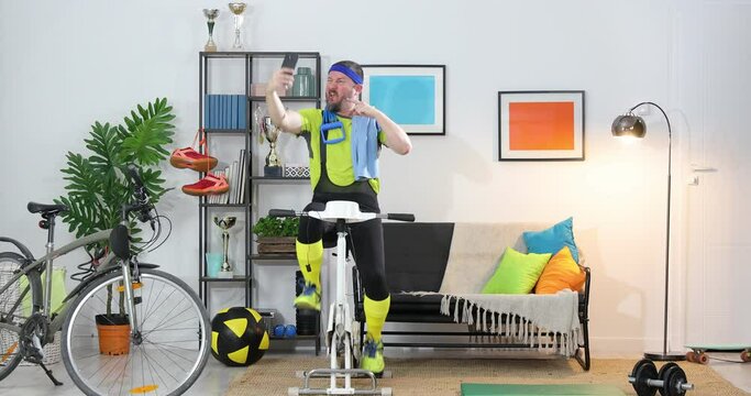funny energetic athlete on exercise bike training at home and taking selfie , Sportsman in retro sportswear recording a cool cycling video on camera in an apartment.