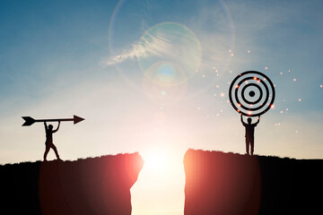 Silhouette men throwing arrow and holding dartboard on mountain with blue sky and sunlight for...