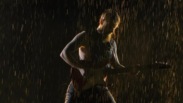 Emotional performance of a rock guitarist in a dark studio in the pouring rain. The musician plays the melody on the electric guitar and shakes his head to the beat of the music. Close up. Slow motion