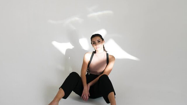 a girl in suspenders and black pants without shoes in makeup and with stylish hair pins on her hair sits on a white background in the studio, and a light flare walks along her