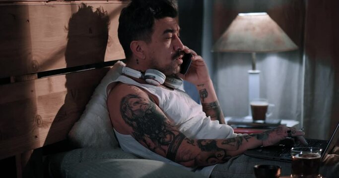Dangerous man with a mustache and tattoos talks on the phone, uses a laptop, smokes a cigar and drink beer.  A man is talking on the phone and using a computer