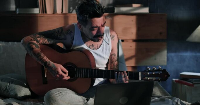 Tattooed man plays a girl on Valentine's Day using a video call on a laptop. The man in the undershirt entertains his love in quarantine by playing guitar online. A man plays guitar in a live stream