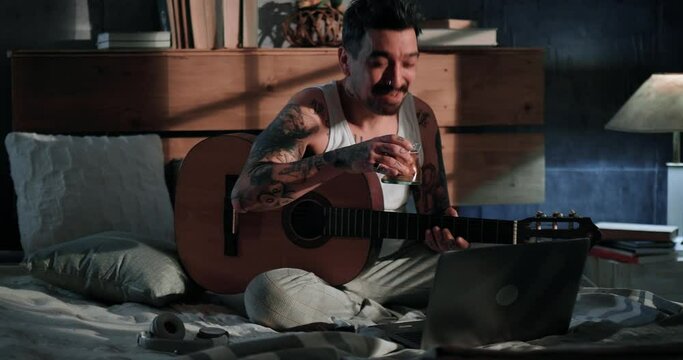 Man with tattoos in isolation toasts in a video call with his girlfriend playing guitar for Valentine's Day. Tattoos all over the body.  Online playing and virtual socializing
