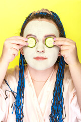 Woman with blue afro braids face in a frozen mask of green clay with slices of cucumbers in front...