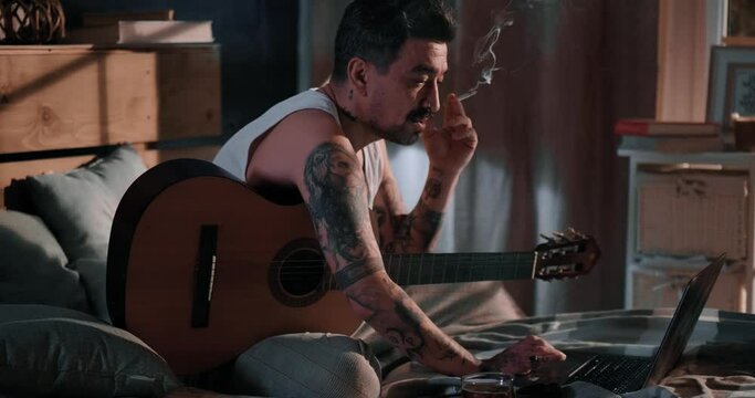 Tattooed man with a guitar starts a video call with a colleague using a video call for an online gig. Due to quarantine, musicians play from home online for people, social distance during quarantine