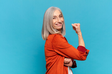 middle age woman feeling happy, satisfied and powerful, flexing fit and muscular biceps, looking...