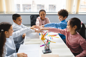 Diverse small schoolchildren putting hands together at classroom