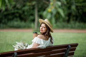 Pregnant young beautiful woman drinks cappuccino in the park. Portrait of a pregnant woman in a white dress and a wicker hat.