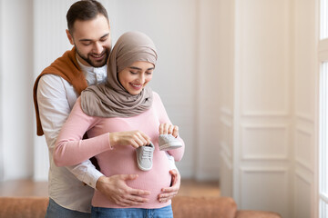 Muslim Husband Holding Tiny Shoes Hugging Pregnant Wife's Belly Indoor