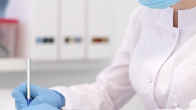 4k woman doctor surgeon in white uniform and mask is writing diagnosis and examination results. Hands in blue gloves close up. Healthcare and medicine
