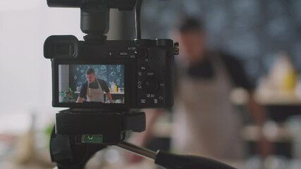 Rack focus shot of male blogger in apron smelling and demonstrating ingredients while recording culinary vlog with professional camera in kitchen