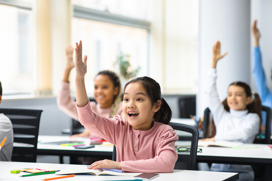Diverse small schoolchildren raising hands at classroom