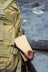 Close up advertising style shot of a green army tactical backpack, a wooden vintage pipe and a notebook on rock steps in the woodlands.