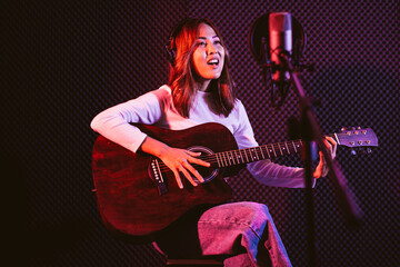 Pretty Asian female singer recording songs by using a studio microphone and pop shield on mic while playing guitar and wearing headphones in the studio. Performance and show in the music business.