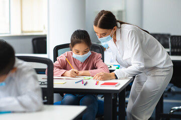 Obraz premium Female teacher in disposable face mask helping small asian schoolgirl