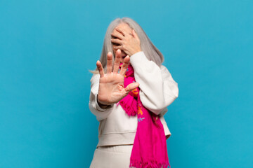 middle age woman covering face with hand and putting other hand up front to stop camera, refusing...