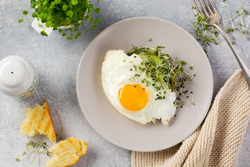Traditional English breakfast with fried eggs with arugula microgreen in gray ceramic plate on gray concrete old background. Top view.