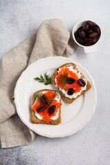 Two sandwiches with smoked pink salmon olives, kalamata, microgreens and cream cheese on gray ceramic plate and trendy concrete background. Traditional Scandinavian toast. Top view.