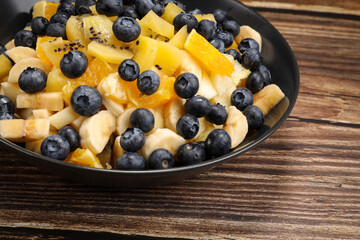 Fruit salad with blueberries, banana, orange, apple and kiwi on a black plate and wooden table. Vitamin cocktail. Summer taste.