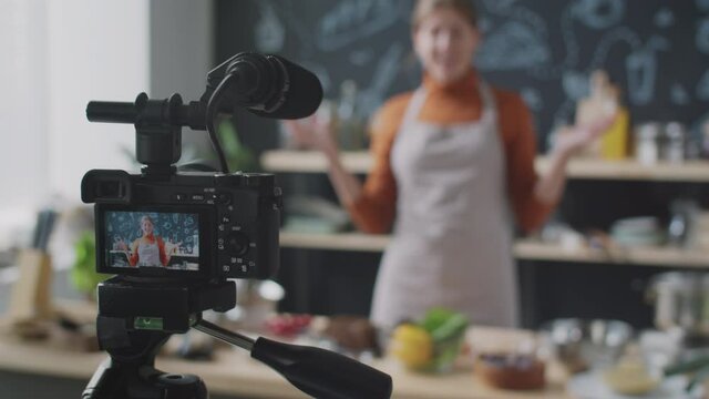 Selective focus shot of professional camera with microphone on tripod recoding video of female food blogger in kitchen