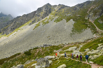 Naklejka premium A group of tourists walk along a mountain trail, among rocky mountains and steep stone slopes in cloudy weather. High Tatras, Slovakia