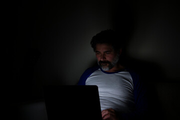 Bearded man sitting on sofa at home at night, working on laptop.