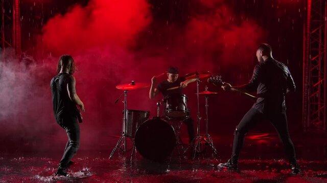 Camera slides over a group of rock musicians who play their instruments with crazy emotions in a dark studio illuminated by red lights. Drum set, electric and bass guitar. Slow motion.