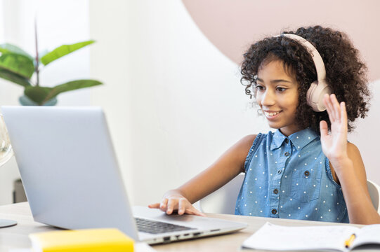 A pretty curly African schoolgirl wearing a headphones using a laptop for studying on the distance, a pretty biracial girl waving into webcam to classmates, to teacher or online tutor on the screen