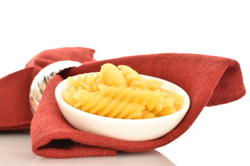 Bright yellow uncooked Fusilli pasta in a white plate on a red linen napkin, close-up, isolated on white.