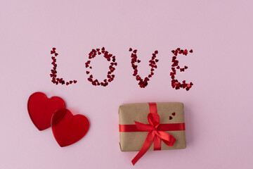 top view of two red transparent hearts, wrapped gift box with red ribbon, inscription love made from confetti hearts on light pink background, copy space, valentines day concept.