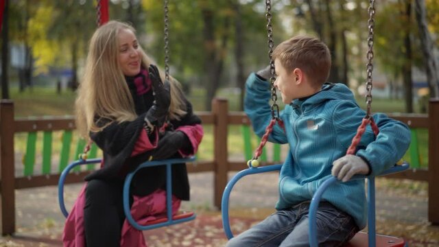Joyful relaxed Caucasian boy sitting on swings talking with mother and giving high-five. Positive boy resting on weekends with beautiful smiling woman. Happy family on playground outdoors.