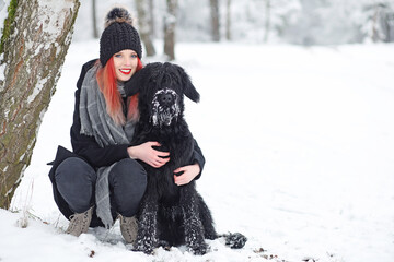 Fototapeta na wymiar Smiling attractive red hair girl is posing with her black Schnauzer dog next to a tree trunk in wintry nature.