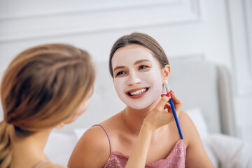 Blonde girl applying facial mask on her smiling friends face