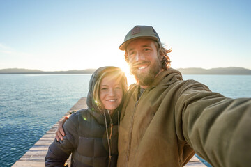 young couple taking selfie while hiking near lake during sunrise