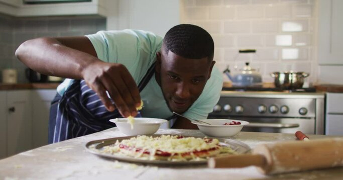 African american man making pizza in kitchen sprinkling grated cheese