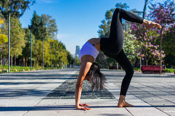 Young girl in grey sportswear, leggings and bra practicing yoga outdoors, brunette girl standing on one leg wheel, Bridge pose, practicing in the park.