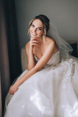 Fototapeta premium A beautiful, sweet, smiling blonde bride in a white lace dress with a long veil sits on an armchair against the background of a wall in a hotel room. Wedding model portrait.