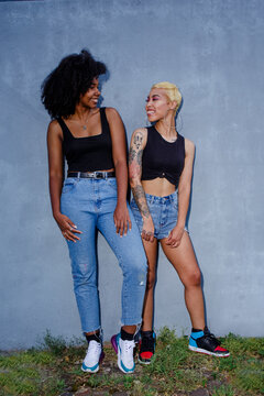 Two smiling friends lean together against a wall in matching clothes