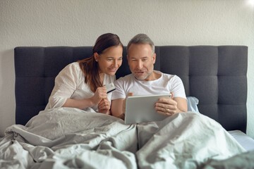 Happy middle-aged couple sitting in bed in the morning drinking coffee and doing online shopping with a digital tablet