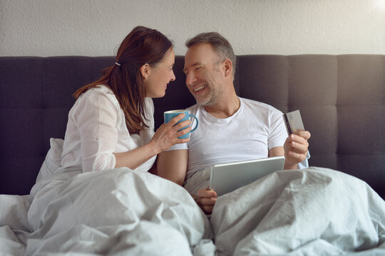 Happy middle-aged couple sitting in bed in the morning drinking coffee and doing online shopping with a digital tablet